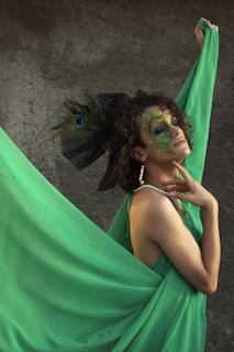 A person poses dramatically with vibrant green fabric draped around them, adorned with elaborate makeup featuring peacock feather designs around the eyes. A large feathered headpiece and pearl jewelry add to the theatrical appearance. The background presents a textured, neutral surface that contrasts with the vivid colors of the ensemble.