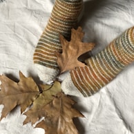 A pair of feet in cozy, striped socks resting on a soft, white fabric, accompanied by dried brown autumn leaves. The socks feature shades of brown, orange, and gray, complementing the earthy tones of the leaves.