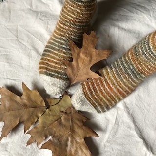 A pair of feet in cozy, striped socks resting on a soft, white fabric, accompanied by dried brown autumn leaves. The socks feature shades of brown, orange, and gray, complementing the earthy tones of the leaves.