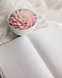 a white cup with a pink flower in it next to an open notebook