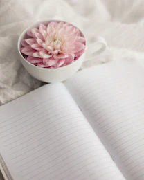 a white cup with a pink flower in it next to an open notebook