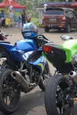 Several motorcycles are parked closely together. The central motorcycle is blue with a matching helmet resting on the seat. Surrounding motorcycles are in black and green colors. In the background, there are people and a brown SUV beside a colorful tent, suggesting a busy street or event scene.
