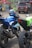 Several motorcycles are parked closely together. The central motorcycle is blue with a matching helmet resting on the seat. Surrounding motorcycles are in black and green colors. In the background, there are people and a brown SUV beside a colorful tent, suggesting a busy street or event scene.