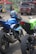 Several motorcycles are parked closely together. The central motorcycle is blue with a matching helmet resting on the seat. Surrounding motorcycles are in black and green colors. In the background, there are people and a brown SUV beside a colorful tent, suggesting a busy street or event scene.