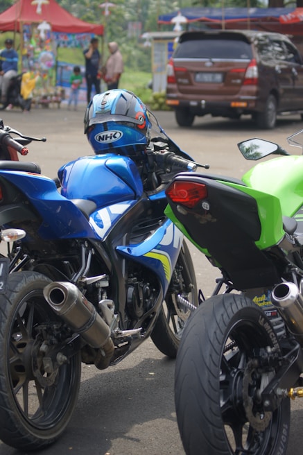 Several motorcycles are parked closely together. The central motorcycle is blue with a matching helmet resting on the seat. Surrounding motorcycles are in black and green colors. In the background, there are people and a brown SUV beside a colorful tent, suggesting a busy street or event scene.