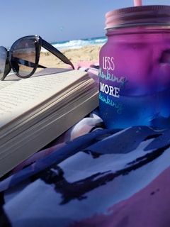 A beach scene with a pair of sunglasses resting on an open book, with partially legible handwriting. The focus is also on a large, colorful insulated bottle that reads 'Less Thinking, More Drinking', set against a sandy beach and ocean waves in the background.