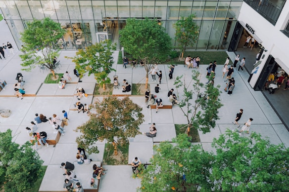 An aerial view of a modern outdoor public space with numerous people scattered around. The area is designed with large concrete tiles and interspersed with trees providing some greenery. People are sitting on benches, walking, or casually lounging around, with a tall building in the background that has large glass windows displaying an interior setup.
