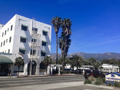A white multi-story building with green awnings and an external staircase is surrounded by palm trees. The background features a clear blue sky and distant mountains. A street with parked cars and a parking sign is visible in the foreground.