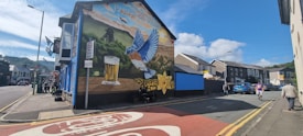 A street scene with a large, vibrant mural painted on the side of a building. The mural features a large bird in flight, a pint of beer, and a yellow daffodil against a backdrop of hills and a coal mine structure. The road is lined with buildings and a few people are walking. The sky is clear with some clouds, and there's a mix of vehicles parked along the street.