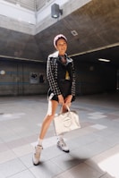 A person stands in a well-lit, industrial-looking space wearing a stylish outfit. The outfit includes a black jacket adorned with metal eyelets, a pair of high-top sneakers, and white socks. They are holding a metallic-colored designer bag and the lighting creates a spotlight effect on them.