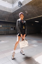 A person stands in a well-lit, industrial-looking space wearing a stylish outfit. The outfit includes a black jacket adorned with metal eyelets, a pair of high-top sneakers, and white socks. They are holding a metallic-colored designer bag and the lighting creates a spotlight effect on them.