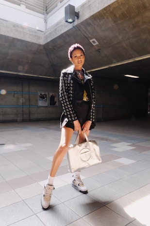A person stands in a well-lit, industrial-looking space wearing a stylish outfit. The outfit includes a black jacket adorned with metal eyelets, a pair of high-top sneakers, and white socks. They are holding a metallic-colored designer bag and the lighting creates a spotlight effect on them.