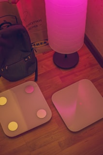 A cozy interior scene featuring a glowing floor lamp casting a pinkish hue over the area. Nearby, a backpack is propped up against a brown paper shopping bag with an IKEA logo. Two square digital bathroom scales with circular accents are placed side by side on a wooden floor.