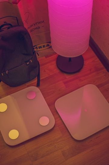 A cozy interior scene featuring a glowing floor lamp casting a pinkish hue over the area. Nearby, a backpack is propped up against a brown paper shopping bag with an IKEA logo. Two square digital bathroom scales with circular accents are placed side by side on a wooden floor.