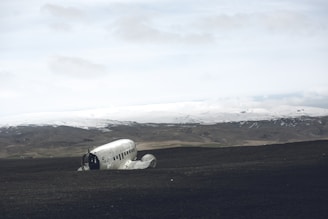 A crashed plane fuselage lies on a desolate, dark sandy landscape. The background features a range of snowy mountains under a cloudy sky, creating a stark contrast between the foreground and the horizon.