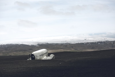 A crashed plane fuselage lies on a desolate, dark sandy landscape. The background features a range of snowy mountains under a cloudy sky, creating a stark contrast between the foreground and the horizon.