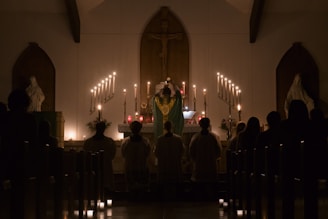 A church interior with a dimly lit altar adorned with numerous candles. A religious figure in a robe stands at the altar, facing a congregation seated in pews. Statues are positioned on either side of the altar, enhancing the sacred ambiance.