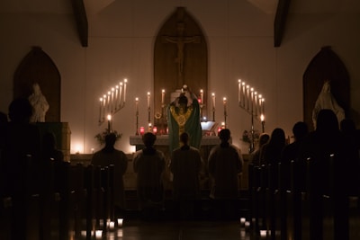 A church interior with a dimly lit altar adorned with numerous candles. A religious figure in a robe stands at the altar, facing a congregation seated in pews. Statues are positioned on either side of the altar, enhancing the sacred ambiance.