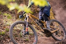 A cyclist is navigating a mountain bike through a forested trail. The bike has a tan frame with the brand logo visible and is equipped with rugged tires suited for off-road terrain. The rider is wearing protective gear including a helmet, gloves, and knee-pads. Surrounding the trail are fallen leaves and branches, which suggest a natural, rugged environment.