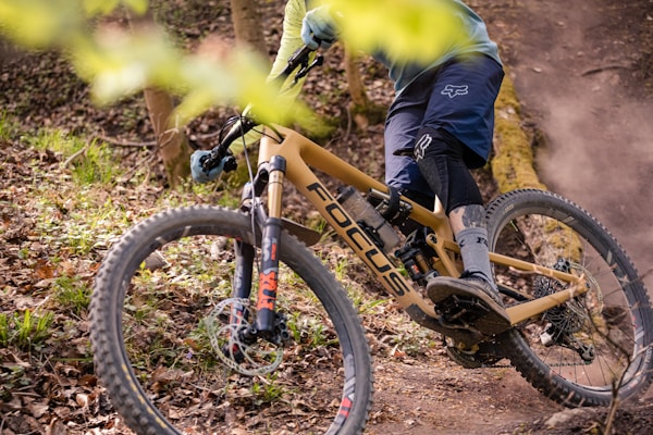 A cyclist is navigating a mountain bike through a forested trail. The bike has a tan frame with the brand logo visible and is equipped with rugged tires suited for off-road terrain. The rider is wearing protective gear including a helmet, gloves, and knee-pads. Surrounding the trail are fallen leaves and branches, which suggest a natural, rugged environment.