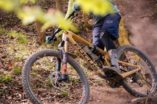 A cyclist is navigating a mountain bike through a forested trail. The bike has a tan frame with the brand logo visible and is equipped with rugged tires suited for off-road terrain. The rider is wearing protective gear including a helmet, gloves, and knee-pads. Surrounding the trail are fallen leaves and branches, which suggest a natural, rugged environment.