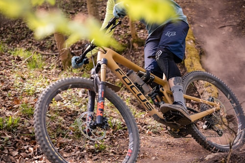 A cyclist is navigating a mountain bike through a forested trail. The bike has a tan frame with the brand logo visible and is equipped with rugged tires suited for off-road terrain. The rider is wearing protective gear including a helmet, gloves, and knee-pads. Surrounding the trail are fallen leaves and branches, which suggest a natural, rugged environment.