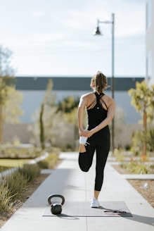 A person is standing on an exercise mat outdoors, performing a standing quad stretch with one hand holding their foot behind them. They are dressed in black athletic wear. In the foreground, a kettlebell and a jump rope are placed on the mat. Surrounding the walkway are plants and a building in the background.