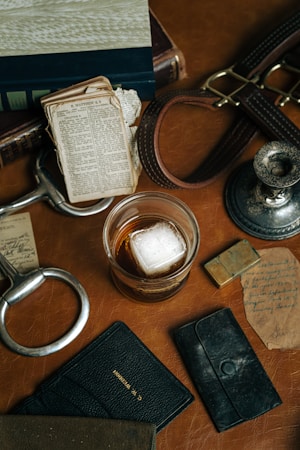 A rustic tabletop is adorned with a collection of vintage items including a glass of whiskey with a large ice cube, aged books, a worn leather strap, an antique candlestick holder, old paper receipts, a black leather wallet, and a metallic bottle opener. The scene evokes a sense of nostalgia and timeless elegance.