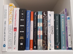 A collection of various books arranged vertically on a white shelf, including titles related to mindfulness, personal development, and self-help. The books have colorful spines and varying thicknesses, creating a visually interesting display.