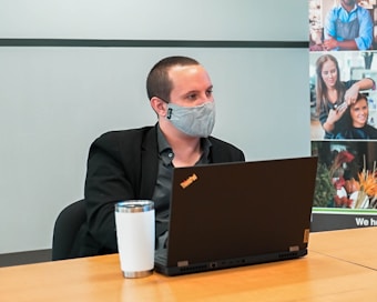 A person is sitting at a conference table wearing a gray face mask and a black jacket. There is a silver and white tumbler and a black laptop with a ThinkPad logo in front of them. The background includes a collage of images showing various professions.