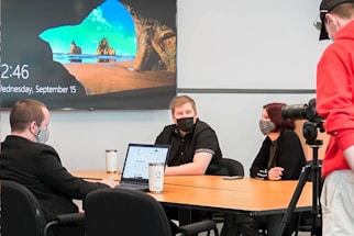 Four people are in a meeting room setting, with one person standing and operating a camera on a tripod. Three individuals are seated at a table, two men and a woman, all wearing masks. A laptop is open on the table in front of them, and a large screen on the wall displays a scenic landscape with a time and date. Coffee cups are also on the table.