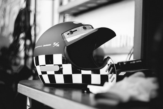 A black and white checkered helmet is placed on a wooden shelf. The helmet has a visor and branding or logos on the side. The background contains blurred shelving and items, giving a sense of an indoor setting.