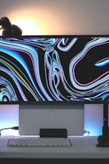 A modern computer setup featuring a large monitor displaying an abstract, colorful swirling pattern. Below the monitor, there is a sleek keyboard and a mouse on a white tabletop, with a backlit effect casting a glow. A black lamp is positioned in the top left corner.