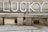 A rustic wooden bench with the word 'LUCKY' painted on the backrest in bold white letters. The wood appears weathered and aged, showing various shades of brown and gray. The setting suggests an outdoor environment, with concrete pavement beneath the bench.