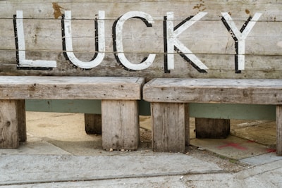 A rustic wooden bench with the word 'LUCKY' painted on the backrest in bold white letters. The wood appears weathered and aged, showing various shades of brown and gray. The setting suggests an outdoor environment, with concrete pavement beneath the bench.