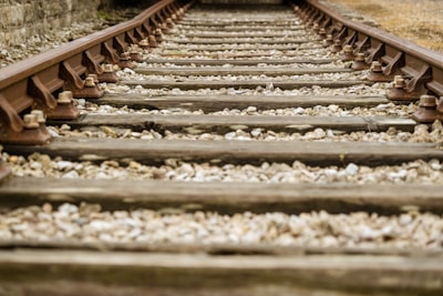 Wooden railroad tracks are laid over gravel, stretching into the distance. The metal rails are secured with bolts, and the track is flanked by grassy and dirt areas, suggesting an outdoor, rustic setting.