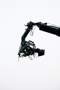 A professional video camera is mounted on a crane arm against a plain, light background. The camera is surrounded by wires and is equipped with various attachments necessary for filming.