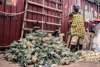 A vendor stands beside a large pile of pineapples, displayed for sale at an outdoor marketplace. The vendor is wearing a colorful apron and standing against a backdrop of a corrugated metal fence. Nearby are wooden planks, and bricks are used as weights to hold down a cloth.
