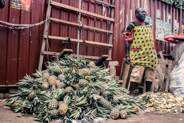 A vendor stands beside a large pile of pineapples, displayed for sale at an outdoor marketplace. The vendor is wearing a colorful apron and standing against a backdrop of a corrugated metal fence. Nearby are wooden planks, and bricks are used as weights to hold down a cloth.