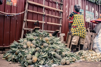 A vendor stands beside a large pile of pineapples, displayed for sale at an outdoor marketplace. The vendor is wearing a colorful apron and standing against a backdrop of a corrugated metal fence. Nearby are wooden planks, and bricks are used as weights to hold down a cloth.