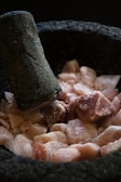 A mortar and pestle with coarse pink Himalayan salt rocks inside. The surface of the mortar and pestle is rough and dark, suggesting it is made of stone. The salt is in irregular chunks, some covered with a light dusting of powder.