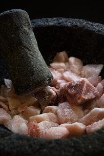 A mortar and pestle with coarse pink Himalayan salt rocks inside. The surface of the mortar and pestle is rough and dark, suggesting it is made of stone. The salt is in irregular chunks, some covered with a light dusting of powder.