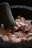 A mortar and pestle with coarse pink Himalayan salt rocks inside. The surface of the mortar and pestle is rough and dark, suggesting it is made of stone. The salt is in irregular chunks, some covered with a light dusting of powder.