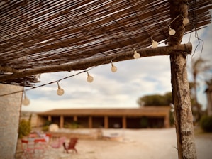 A rustic outdoor setting featuring a wooden pergola with string lights. The foreground shows details of the pergola's wooden beams and bamboo covering. In the background, blurred elements include some red chairs and tables on a stone patio and a building with arches.