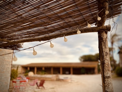 A rustic outdoor setting featuring a wooden pergola with string lights. The foreground shows details of the pergola's wooden beams and bamboo covering. In the background, blurred elements include some red chairs and tables on a stone patio and a building with arches.