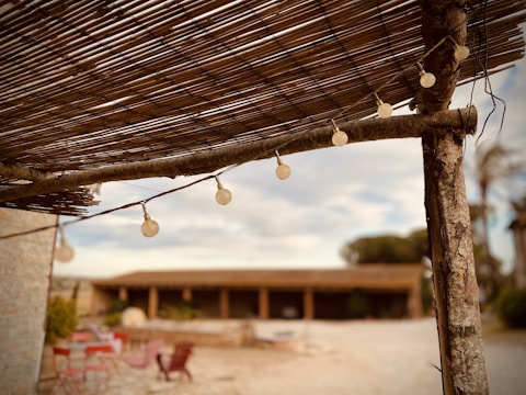 A rustic outdoor setting featuring a wooden pergola with string lights. The foreground shows details of the pergola's wooden beams and bamboo covering. In the background, blurred elements include some red chairs and tables on a stone patio and a building with arches.