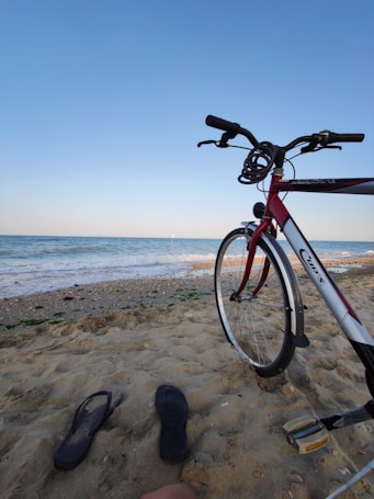 A sandy beach scene with a bicycle resting on the sand. The ocean extends into the horizon under a clear blue sky. Two flip-flops are lying nearby on the sand with some seaweed scattered along the shore.
