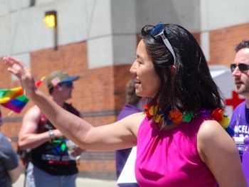 A woman wearing a vibrant pink top and colorful lei is smiling and waving. She has sunglasses perched on her head. In the background, others are present, with one person holding a rainbow flag, suggesting a festive or parade-like atmosphere.