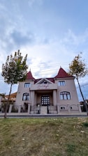 A two-story building with beige walls and red-tiled roofs, featuring symmetrical architecture typical of a restaurant. The entrance is grand, with steps leading to a double door under an awning that reads 'To Chef Restaurant'. It is flanked by two small trees and surrounded by a well-kept lawn, enclosed by a black iron fence. The sky overhead is cloudy, creating a tranquil and picturesque setting.