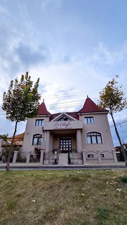 A two-story building with beige walls and red-tiled roofs, featuring symmetrical architecture typical of a restaurant. The entrance is grand, with steps leading to a double door under an awning that reads 'To Chef Restaurant'. It is flanked by two small trees and surrounded by a well-kept lawn, enclosed by a black iron fence. The sky overhead is cloudy, creating a tranquil and picturesque setting.